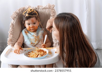 Unhappy mixed race toddler child messy tomato sauce on face crying with mother sitting together beside in the room. Upset messy little girl mix race hungry eating spaghetti with mom at home. Lifestyle