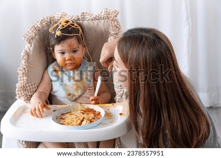 Unhappy mixed race toddler child messy tomato sauce on face crying with mother sitting together beside in the room. Upset messy little girl mix race hungry eating spaghetti with mom at home. Lifestyle