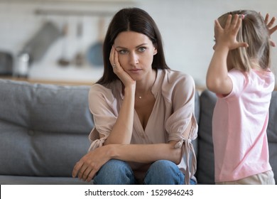 Unhappy mother having problem with noisy naughty little daughter screaming, demanding attention, child tantrum manipulation concept, tired thoughtful mum holding head, sitting on couch at home