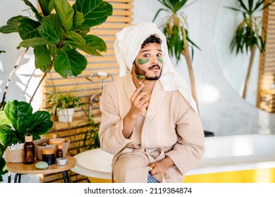 Unhappy multiethnic man feels disgruntled with consternation facial expression in fresh bathrobe, towel and green eye patches using jade roller looking in camera. Male portrait in bathroom interior. - Shutterstock ID 2119384874