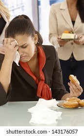 Unhappy office worker sitting at desk, crying, eating doughnut.
