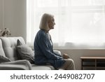 Unhappy older woman sit on sofa in living room, feels lonely, think over health problems, suffers from loneliness. Faced up with loss or illness disorder, having depression or apathy, melancholic mood