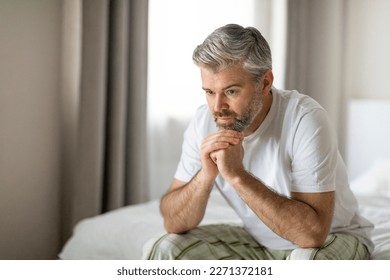 Unhappy pensive middle aged grey-haired handsome man wearing pajamas homewear sitting on bed alone at home, suffering from depression, loneliness or anxiety, copy space. Permacrisis, midlife crisis - Powered by Shutterstock