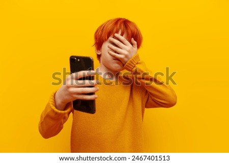 unhappy red-haired child boy using smartphone and showing facepalm on yellow isolated background, confused teenager with orange hairstyle loses online and makes mistakes