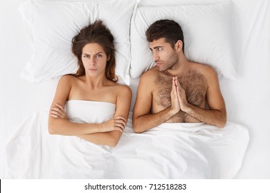 Unhappy Regretful Bearded Man Lying In Bed With His Wife And Holding Hands Pressed Together, Feeling Sorry, Asking Her To Forgive Him, Woman Having Angry Mad Expression, Keeping Arms Crossed