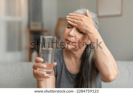 Unhappy sad caucasian old lady hold glass of water and touching forehead, suffering from fever, menopause in living room interior. Health problems, treatment of disease, high pressure and migraine