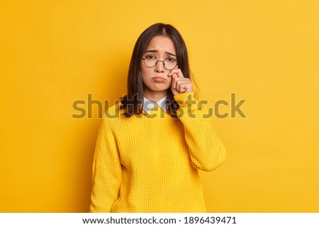 Unhappy sad dejected woman with eastern appearance rubs tears wants to cry feels desperate has problems in life wears round spectacles and casual jumper isolated over vivid yellow background.