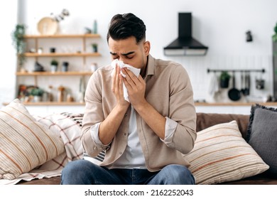 Unhappy sad indian or arabian guy suffering from fever and flu, sitting on sofa in living room, blowing nose and sneezing in napkin, need treatment of illness, and doctors consultation