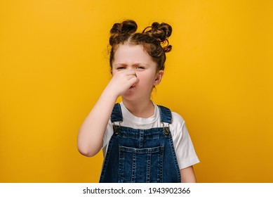 Unhappy Sad Little Preschool Girl Kid Feeling Bad And Dislike Smell And Hold Nose From Funny Odor, Dressed In White T-shirt, Isolated On Yellow Studio Background, Six Year Child Looking At Camera