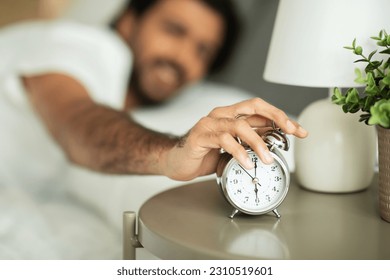 Unhappy sad millennial middle eastern guy turns off alarm clock, lies on bed in bedroom interior, blurred, close up. Wake up early, time to work, sleep problems, ad and offer - Shutterstock ID 2310519601
