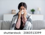 Unhappy sad young indian female in plaid suffering from fever and flu on sofa, blowing nose in napkin in living room interior. Covid-19 lockdown, treatment of illness, cold and runny, copy space