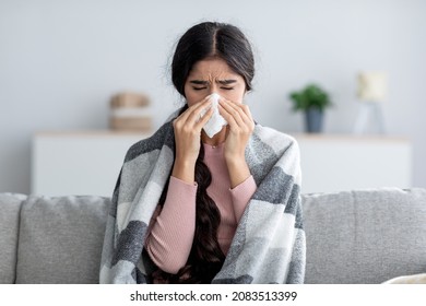 Unhappy sad young indian female in plaid suffering from fever and flu on sofa, blowing nose in napkin in living room interior. Covid-19 lockdown, treatment of illness, cold and runny, copy space - Shutterstock ID 2083513399