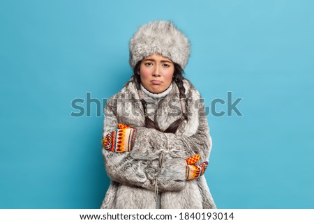Unhappy scandinavian woman in fur hat and coat crosses hands and feels freezing trembles during severe frosty day wears winter outerwear. Yakut girl shivers from cold poses against blue background