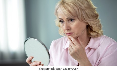 Unhappy Senior Female Looking At Sagging Skin Face In Mirror, Old Age Appearance