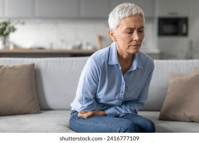 Unhappy Senior Woman Having Stomachache Suffering From Abdominal Pain Sitting On Sofa At Home. Mature Lady Having Painful Spasm Touching Belly. Stomach Diseases In Older Age Concept - Powered by Shutterstock