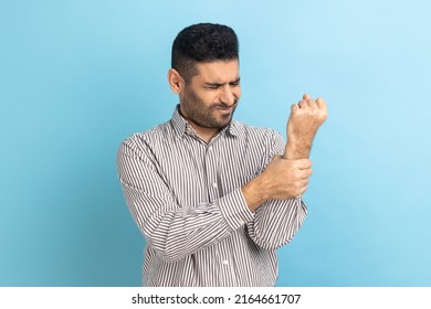 Unhappy Sick Bearded Businessman Touching Painful Hand, Suffering Trauma, Sprain Wrist, Feeling Ache Of Carpal Tunnel Syndrome, Wearing Striped Shirt. Indoor Studio Shot Isolated On Blue Background.