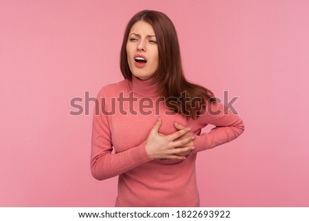 Unhappy sick brunette woman holding hands on chest feeling acute pain in chest, risk of stroke, heart attack. Indoor studio shot isolated on pink background