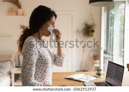 Unhappy sick woman, adult student, remote employee staying at home due to flu infection, suffering from snuffles, seasonal allergy, blowing runny nose, covering face with paper tissue