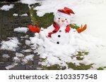 Unhappy snowman in mittens, red scarf and cap is melting  outdoors in sunlight on snowy background mixed with green grass and wet pavement. Approaching spring, warm winter, climate change concept