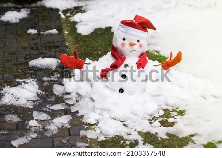 Unhappy snowman in mittens, red scarf and cap is melting  outdoors in sunlight on snowy background mixed with green grass and wet pavement. Approaching spring, warm winter, climate change concept