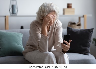 Unhappy Stressed Mature Middle Aged Woman Looking At Phone Screen, Sitting On Couch In Living Room, Upset Frustrated Senior Female Reading Bad News In Message Or Social Network, Problem With Device