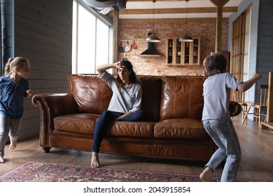 Unhappy Stressed Young Mother Sitting On Sofa, Feeling Exhausted Of Hyperactive Small Children Running Playing Around, Needs Rest. Joyful Little Kids Irritating Tired Mum, Upbringing Problems Concept.