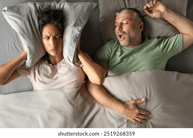 Unhappy tired woman covering her ears with pillow, can not handle loud snore of her husband, top view shot - Powered by Shutterstock