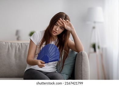 Unhappy Tired Young Caucasian Lady Suffering From Heat, Fever And Headache Touching Forehead Blowing Fan At Herself Sit On Sofa In Living Room Interior. Too Hot, Summer Weather, Overheating At Home