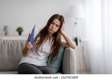 Unhappy Tired Young European Female Suffering From Heat Waving Fan At Herself Sitting On Sofa And Blowing Lips In Living Room Interior. Summer Weather And Overheating Without Air Conditioning At Home