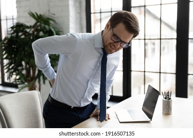 Unhappy Unwell Male Employee Suffer Sitting In Incorrect Posture At Desk In Office, Have Backache Or Muscular Spasm. Unhealthy Man Worker Struggle With Back Muscle Strain. Sedentary Life Concept.