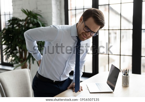 Unhappy unwell male employee suffer sitting in
incorrect posture at desk in office, have backache or muscular
spasm. Unhealthy man worker struggle with back muscle strain.
Sedentary life concept.