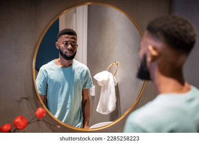 Unhappy upset 30s African American man looking in mirror, standing in bathroom at home, sad depressed black guy feeling dissatisfied with his appearance. Midlife crisis in men - Powered by Shutterstock