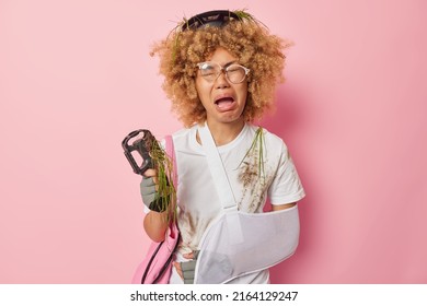 Unhappy woman got injured after falling off bike wears sling cries from despair and painful feelings holds bicyclepedal dressed in white t shirt smeared with dirt isolatd over pink background