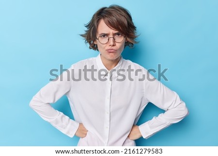 Unhappy woman keeps hands on waist has sulking facial expression wears round spectacles white shirt poses against blue background being angry on someone cringes from awful content judges something