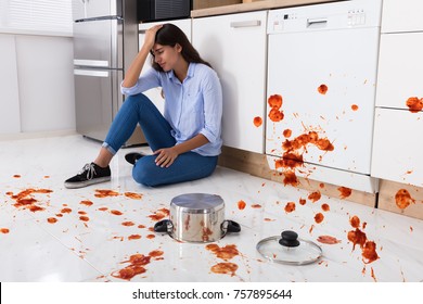 Unhappy Woman Sitting On Kitchen Floor With Spilled Food In Kitchen