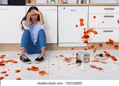 Unhappy Woman Sitting On Kitchen Floor With Spilled Food In Kitchen