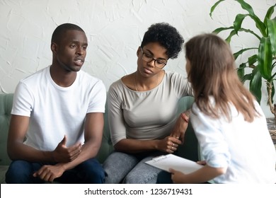 Unhappy young African American couple visiting psychologist, upset man talking about relationships problem with tired frustrated woman, family therapy session, husband and wife sit on couch together