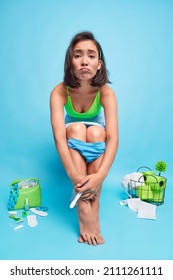 Unhappy young Asian woman poses on toilet bowl with pregnancy test waits for result has panties pulled down on legs surrounded by hygiene items isolated over blue background. Fertility concept