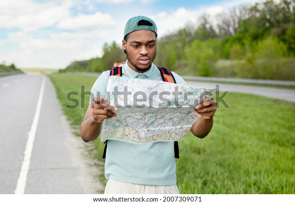 Unhappy young black man hitchhiking on road,\
looking at map, feeling lost, traveling alone by autostop.\
Millennial African American guy standing on highway, cannot find\
his tourist destination