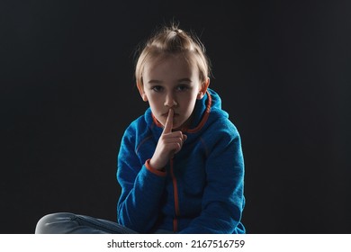 Unhappy Young Boy With A Finger On Lips. Sad Afraid Child Sitting Quiet In Dark. Young Boy Suffering From Anxiety. Stressed Boy Over Black Background.