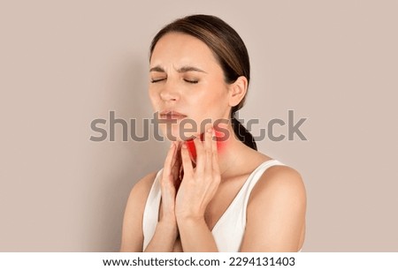Unhappy young brunette woman with closed eyes wearing white top suffering from pain in throat, touching red inflamed zone on her neck, panorama with empty space, beige studio background