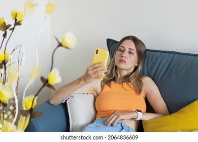 Unhappy Young Caucasian Blonde Woman Relaxing At Home Alone, Sitting On Couch In Comfortable Pose, Blogger Sharing Good News At Social Media Via Mobile Phone. Smiling Woman Enjoying Weekend Order