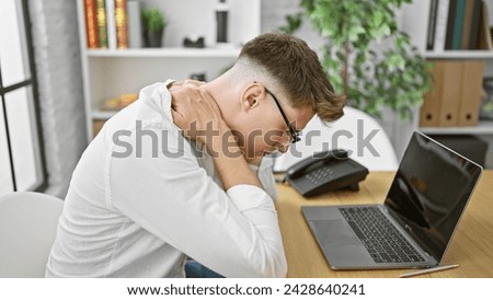 Unhappy young caucasian guy, a business professional, seriously suffering from neck pain at his office, work woes outpouring as he tries to focus on the laptop