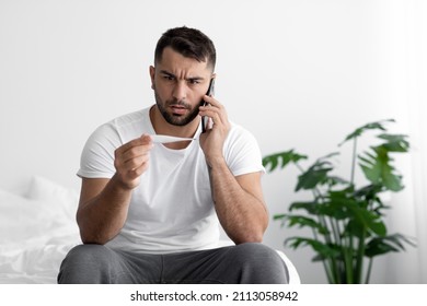 Unhappy Young Caucasian Man Sits On White Bed And Looks At Thermometer With High Temperature, Call By Phone In Bedroom Interior. Sickness, Flu And Cold, Health Problem And Fever. Medical Consultation