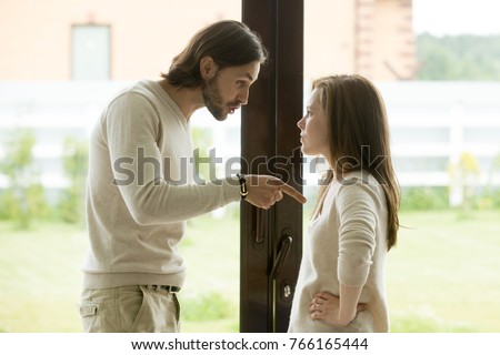 Unhappy young couple arguing standing at house door, angry husband pointing at wife blaming her of problems, conflicts in marriage, bad relationships, man and woman having quarrel or disagreement