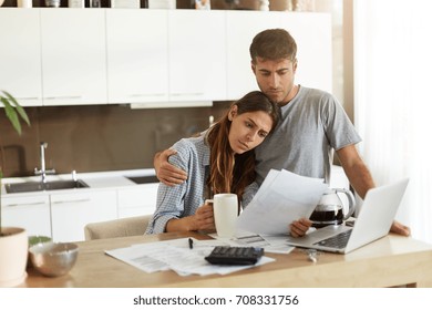 Unhappy Young European Family Facing Financial Troubles: Sad Husband Deep In Thoughts Hugging His Worried Wife Who Is Studying Notification From Bank In Her Hands While Doing Finances In Kitchen