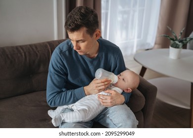 Unhappy Young Father Feeding Newborn Baby With Milk Bottle On Couch At Home. Depressed Single Dad Tired From Sleepless Night, Fatherhood, Take Care Of Infant Sun. Paternity And Paternity Leave Concept