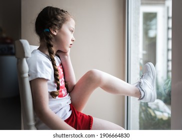 Unhappy Young Girl With Hair Platted Bored In Pandemic Lockdown Sat In Home Stuck Indoors On Chair Looking Out Window Shorts Trainers T-shirt Staring Outside Wanting To Go Outdoors In Sunshine To Play