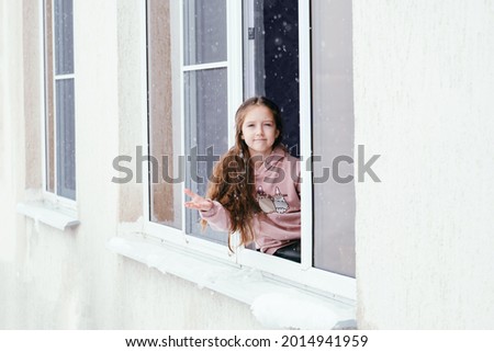 Unhappy young girl sitting on a window sill in open window and feel sad about snowy weather