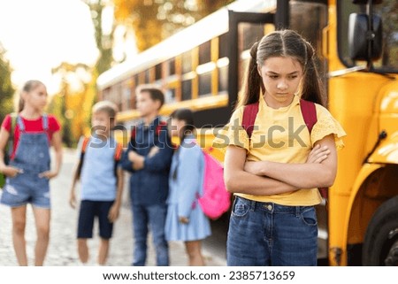 Unhappy young girl standing by herself next to yellow school bus, arms crossed and scowling while other kids laughing and talking behind her, female kid feeling left out and experiencing loneliness